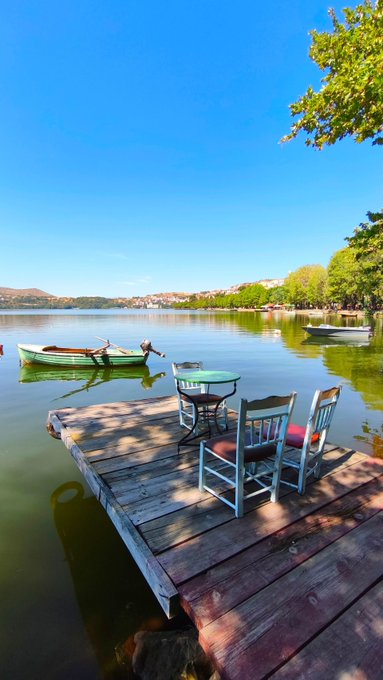 A serene lake scene featuring a wooden dock with a green table and two chairs, with small boats moored nearby and trees casting shadows.
