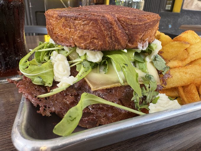 A towering gourmet burger featuring a thick, juicy patty topped with melted cheese, fresh arugula, and creamy cheese spreads, sandwiched between slices of golden toasted bread, served alongside crispy golden fries on a metal tray. The hearty, indulgent presentation fits the "Rate my burger" post, showcasing a restaurant-quality meal ready for review.