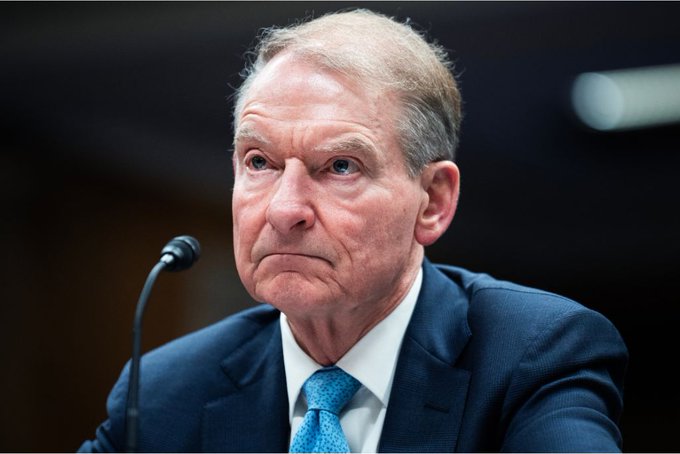 A close-up photograph of an elderly man in a dark suit and light blue tie sitting in front of a microphone with a serious expression.