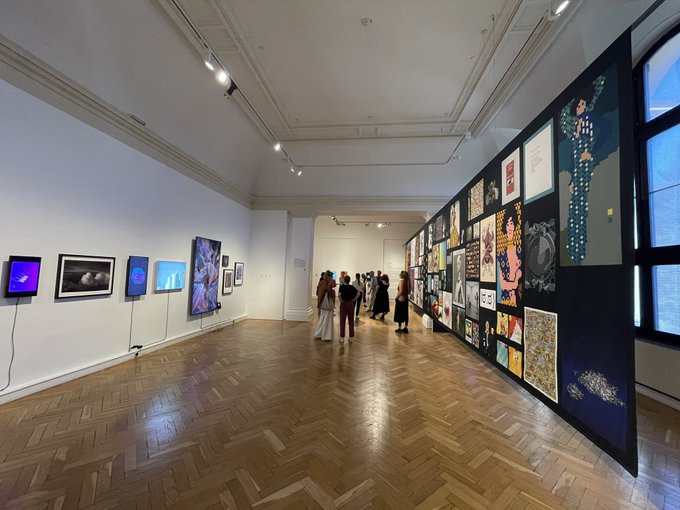 An interior view of an art gallery with herringbone wood floors, white walls displaying framed artworks, and visitors viewing the exhibitions.