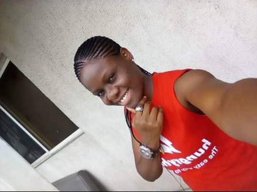The image features a young woman with braids, wearing an orange shirt that reads "doginal dogs". She has her hand on her face and is smiling at the camera. The background of the photo shows a white wall, which contrasts with the vibrant colors of the woman's outfit. There are no other objects or people in the image besides the woman herself.