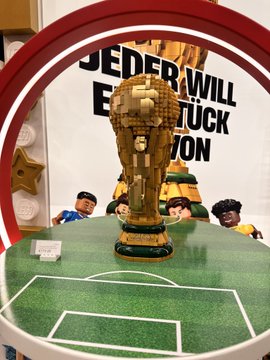 A Lego FIFA World Cup trophy displayed on a miniature soccer field with figures, positioned in front of a sign reading 'JEDER WILL EINE...'