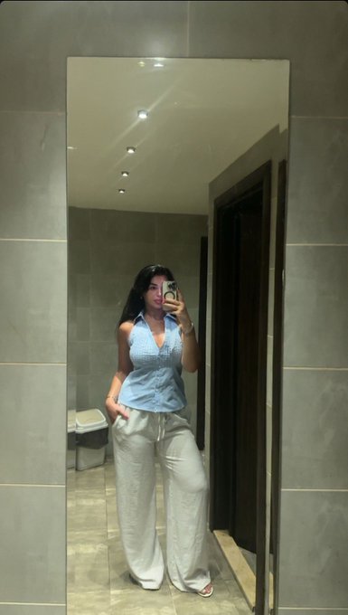 A mirror selfie shows a woman wearing a light blue halter top and white pants, taking a photo with her phone in what appears to be a tiled bathroom or hallway.