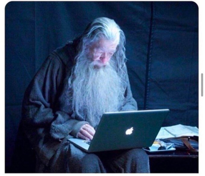 The image features a man with long white hair sitting down, wearing a robe or hooded garment, and using an Apple laptop computer. The background of the image is dark blue, which contrasts with the light-colored man's attire. There are no other objects in the scene besides the man and his laptop.