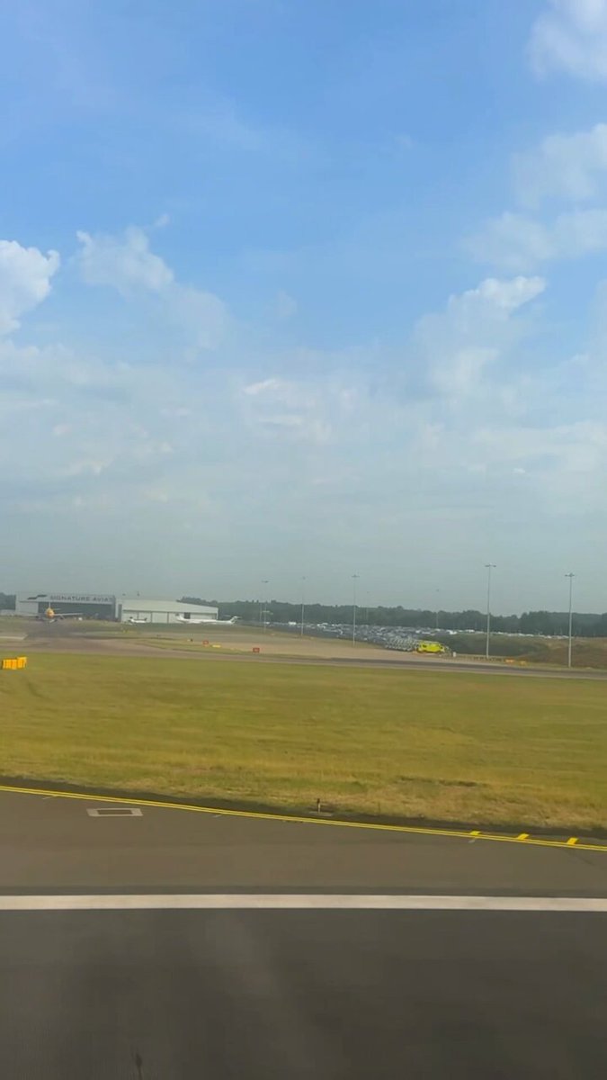 The image shows an airport runway with several airplanes parked along the tarmac. The sky above the runway has a few clouds scattered throughout. In the background, there are buildings that appear to be part of the airport infrastructure. A yellow line runs down the center of the road in front of the runway, and a car is visible on the road as well.