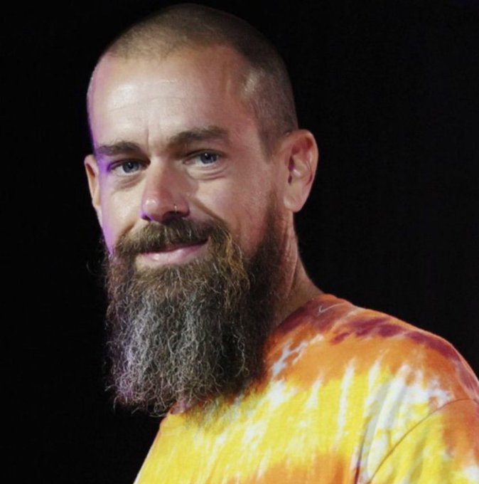A close-up portrait of a bald man with a long brown beard, wearing a yellow and orange tie-dye shirt against a black background.