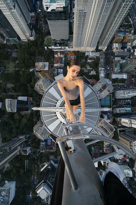 A vertigo-inducing aerial photograph of a woman perched at the edge of a skyscraper's antenna spire, looking down at a sprawling cityscape far below.