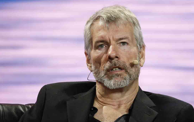 Michael Saylor speaking at an event with a headset microphone, illustrating a post that humorously highlights his shifting messaging about Bitcoin selling strategy from 2020 to 2026.