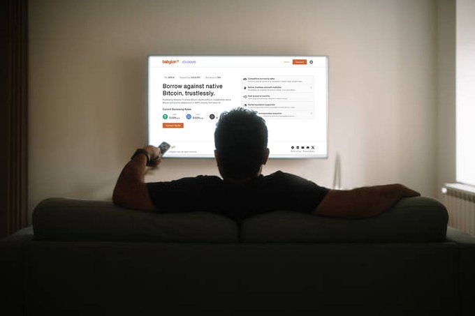 A person viewed from behind relaxes on a couch while watching a TV displaying the Babylon by busdave Bitcoin website, with text "Borrow against native Bitcoin, trustlessly."