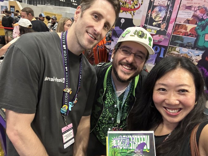 Three smiling people posing together at what appears to be a comic convention, with the man on the left wearing a lanyard that reads 'BRAINSTORM! KEEGAN CON 2024'.
