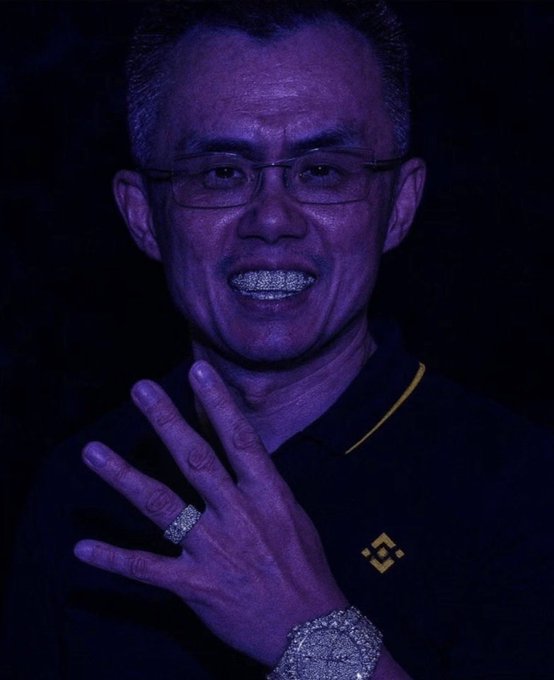 A portrait of a man wearing glasses and a black polo shirt with a yellow logo, displaying diamond grillz and jewelry in purple lighting.