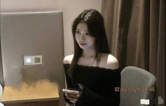 The image shows a young woman with long dark hair wearing a black off-the-shoulder top, smiling at her phone in a hotel room with a timestamp of "02/02/2025 04:45" — likely a screenshot shared in response to the post asking followers to identify the attractive person pictured.