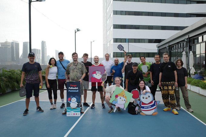 A group photo of approximately 15 people posing on a blue and green pickleball court, with a city skyline in the background; some hold paddles and cardboard cutouts of cartoon characters including a penguin holding a 'Palladium Pickleball' sign.