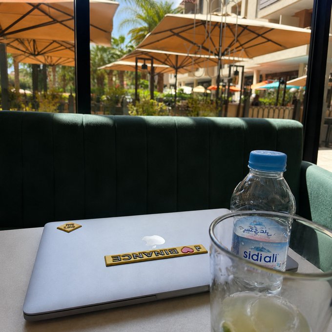 View from a cafe table showing a silver laptop with 'BINANCE' stickers, a bottle of Sidi Ali water, and an empty glass, with outdoor umbrellas visible through the window.