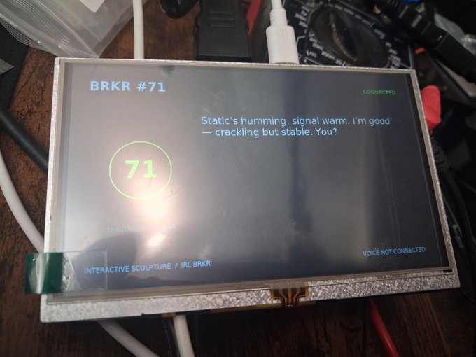 A small electronic display screen shows 'BRKR #71' and a message reading 'Static's humming, signal warm. I'm good — crackling but stable. You?' along with 'INTERACTIVE SCULPTURE / IRL BRKR' and 'VOICE NOT CONNECTED'.
