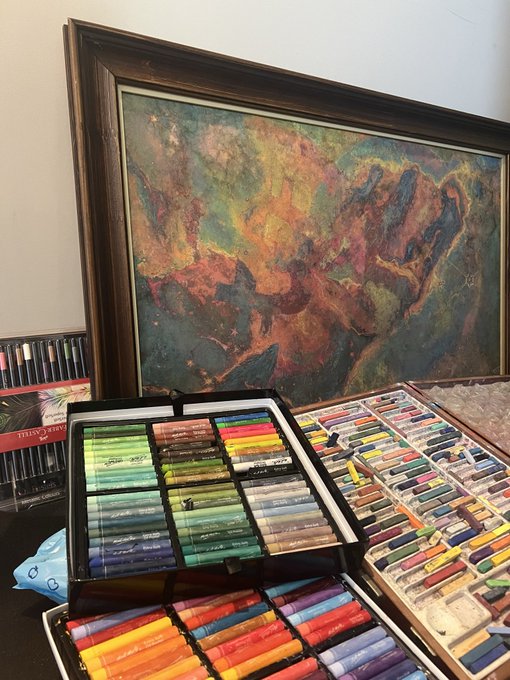 The image features a table with an assortment of art supplies, including various tubes of paint arranged in different colors. The supplies are organized into multiple drawers or compartments, showcasing the artist's collection. In addition to the paints, there are also some pencils and other drawing tools present on the table. A large painting can be seen behind these supplies, adding an artistic touch to the scene.