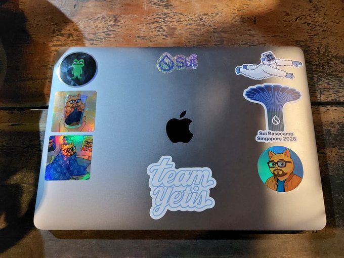 A silver MacBook laptop on a wooden surface decorated with stickers including 'Sui', 'team yetis', 'Sui Basecamp Singapore 2026', and various illustrated cat characters.