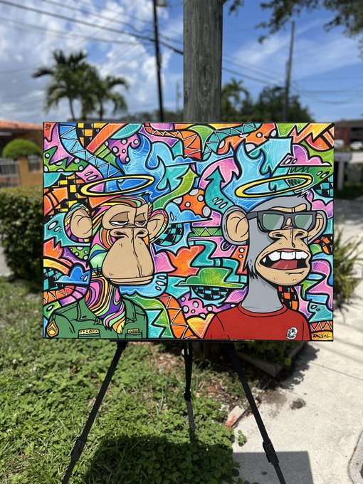 A colorful painting depicting two cartoon monkeys with halos, one wearing sunglasses and a red shirt, sits on an easel outdoors with a blue sky background.