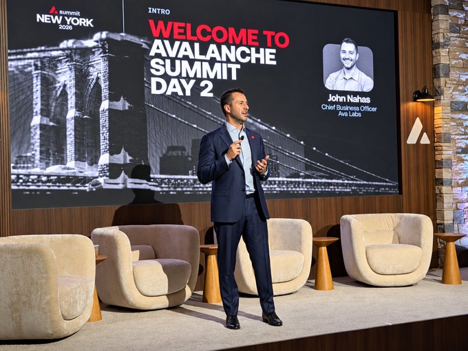A man in a navy suit speaks on stage at the Avalanche Summit Day 2, with a screen behind him reading 'WELCOME TO AVALANCHE SUMMIT DAY 2' and introducing 'John Nahas, Chief Business Officer, Ava Labs' at 'Summit NEW YORK 2025.'