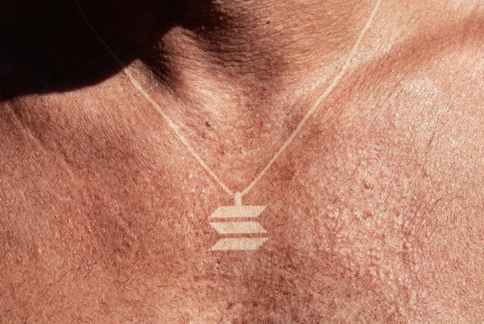 A close-up photo of a gold necklace featuring the 'RIYE' (IVE) logo pendant resting on someone's chest in sunlight.