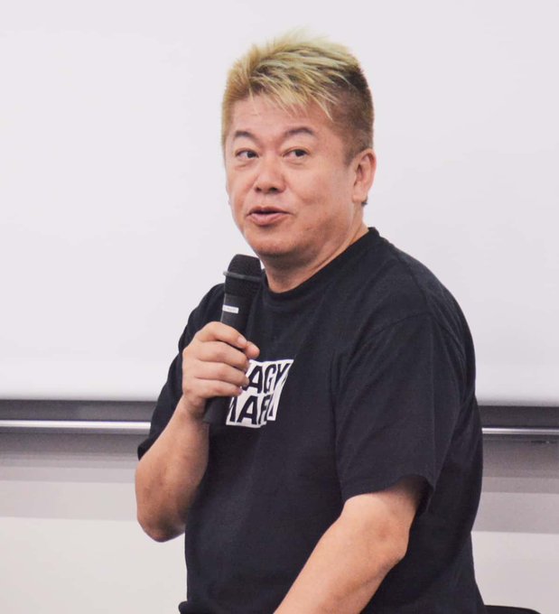 A man with blonde-tipped hair speaks into a microphone while wearing a black "Angry Japan" t-shirt, appearing to be Takafumi Horie as referenced in the SANAE TOKEN post criticizing him.