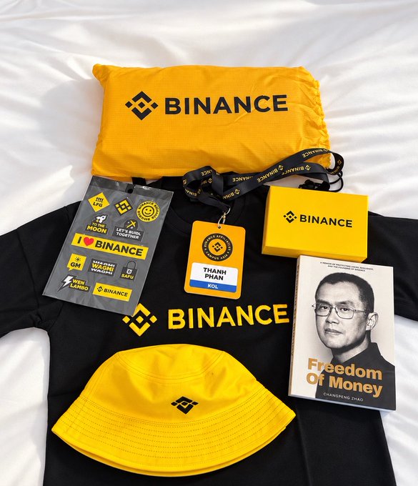 A collection of Binance-branded merchandise laid out on a bed, including a yellow bag, a black hoodie, a yellow bucket hat, a sticker sheet, a name tag reading 'THANH PHAN', and a book titled 'Freedom Of Money' by Changpeng Zhao.