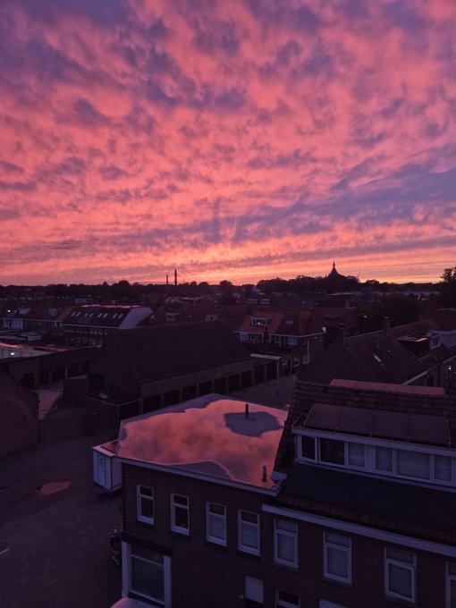 A scenic sunset photograph showing a dramatic pink and orange sky over a residential neighborhood with rooftops, solar panels, and a distant pagoda silhouette.