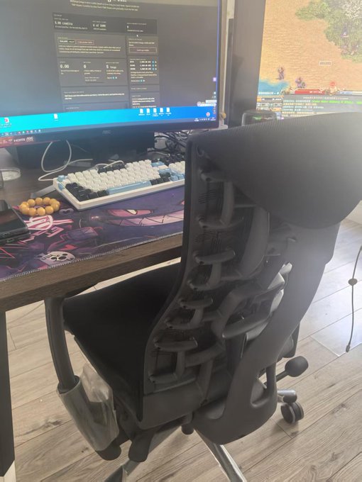 A black mesh ergonomic office chair positioned in front of a desk with a white mechanical keyboard, a monitor displaying system stats, and a game running on a second screen.