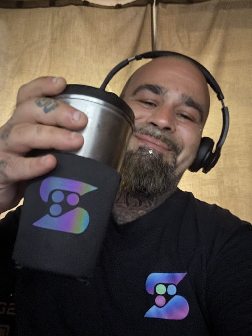 A bearded, tattooed man with headphones around his neck holds up a stainless steel tumbler featuring an iridescent "B" logo, wearing a matching black t-shirt with the same design. The celebratory "MASSIVE WEEK" caption aligns with his wide, triumphant smile, suggesting he's toasting to a successful milestone for the brand or project represented by the logo.