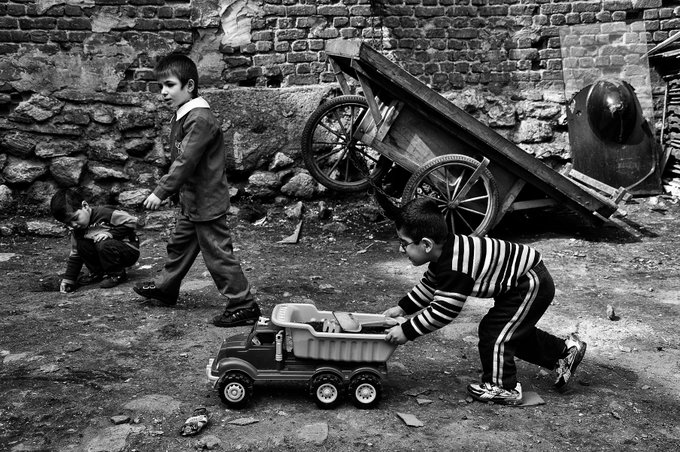 A black and white photograph of three young boys playing in a dirt yard against a stone wall, with one pushing a toy truck while another walks by and a third crouches nearby.