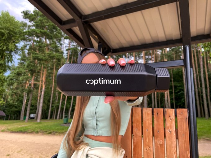 A person with long hair, dressed in a light green outfit and black cap, holds a black "optimum" branded case in front of their face under a wooden shelter, set against a backdrop of tall pine trees. The image aligns with the post's theme of Optimum's growing presence across various infrastructure-focused events and discussions in the crypto and trading space.
