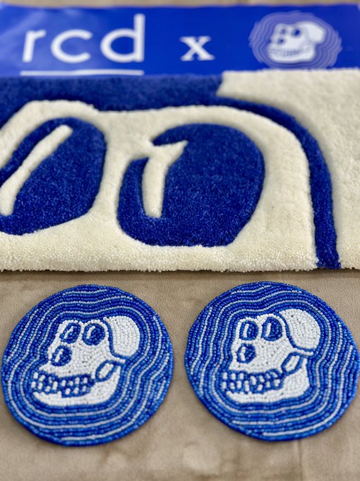 A blue and white "rcd x" tufted rug featuring an ape face design sits above two matching circular embroidered coasters, both displaying the iconic ApeCoin skull logo. The post expresses gratitude to a collector named Glenn for acquiring this collectible keyboard rug and coaster set, highlighting the real-life ("irl") community connections formed through collecting.