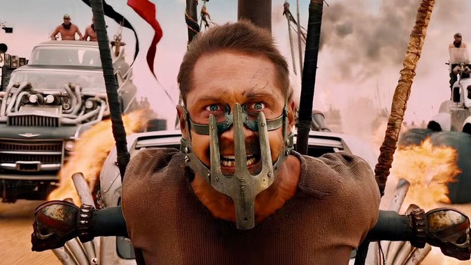 A scene from Mad Max: Fury Road showing the character Nux strapped to the front of a vehicle with an intense expression, set against a fiery post-apocalyptic backdrop.