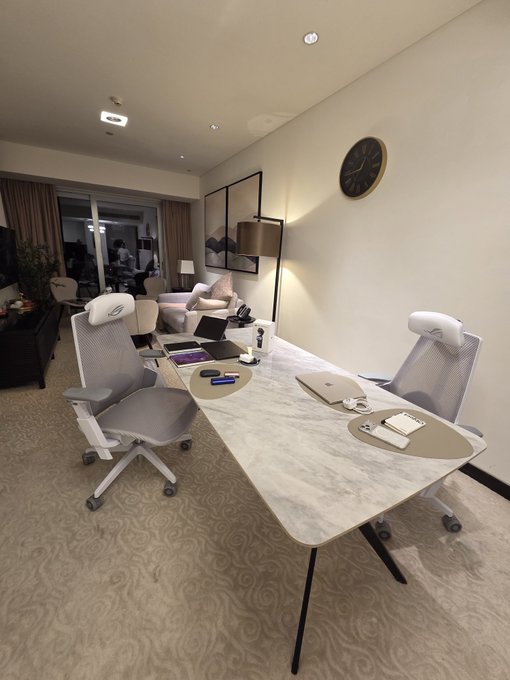 A modern hotel room or office space featuring a white marble desk with two ergonomic mesh chairs, a laptop, a wall clock, and framed artwork in a warmly lit interior.