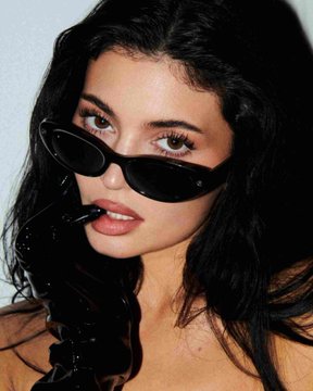 The image features a woman wearing black sunglasses and holding a pair of black gloves. The woman has long hair and is posing for the camera, looking directly at the viewer. There are no texts or other objects present in the image.