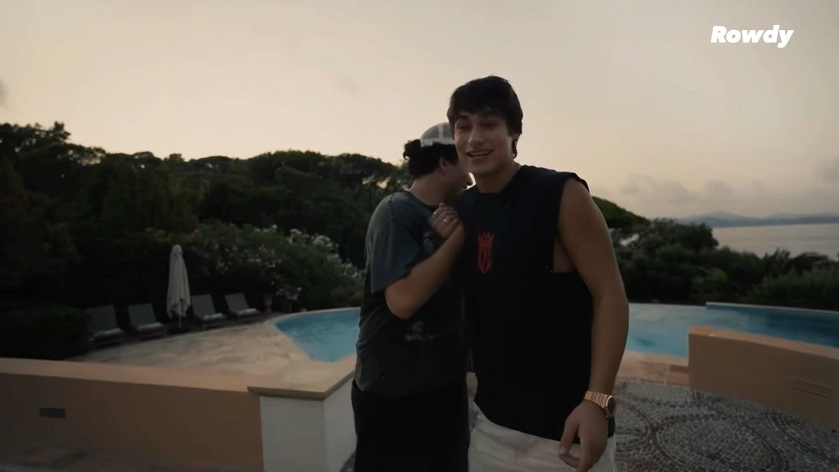 Two young men stand by an infinity pool overlooking water and mountains at dusk, with the watermark 'Rowdy' in the top right corner; one wears a black sleeveless shirt and a gold watch.