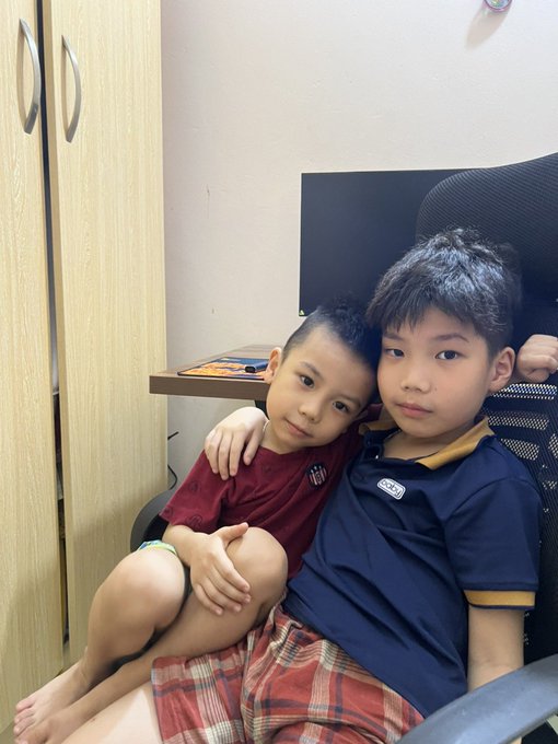 Two young boys pose together on a chair, with the older boy's arm around the younger one, in what appears to be a bedroom with a wardrobe in the background.