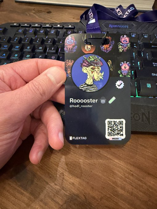 A hand holds a 'FLEXTAG' badge/event pass labeled 'Rooooster' (@hodl_rooster) featuring cartoon NFT character artwork, held above a Redragon keyboard on a wooden desk.