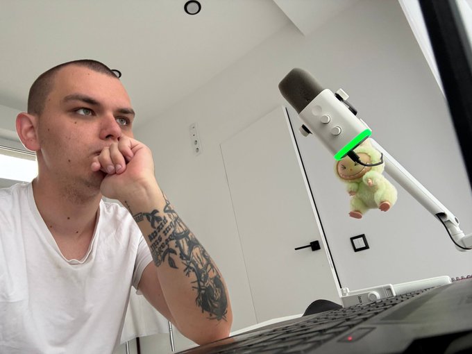 A man with a tattooed arm resting his chin on his hand, looking thoughtfully at a white microphone with a small plush toy hanging from it.