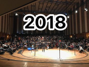 The image shows a large crowd of people gathered inside a building for a conference or event. The room has a stage set up at one end with a microphone, suggesting that there might be a speaker addressing the audience. The year "2018" is prominently displayed in white text above the scene, indicating it's an important date related to the event.