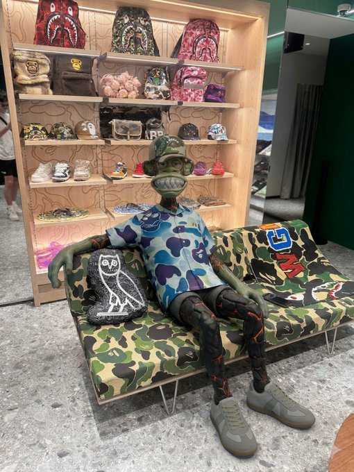 A green-skinned figure wearing a blue camouflage BAPE t-shirt and cap sits on a matching BAPE camo couch, surrounded by shelves of BAPE hats, bags, and accessories, fittingly illustrating the "GM (B)APES" caption.