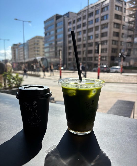 Two iced matcha drinks sit on a dark outdoor table against a sunny urban backdrop with buildings and pedestrians; one is in a black branded reusable cup and the other in a clear plastic cup with a black straw.