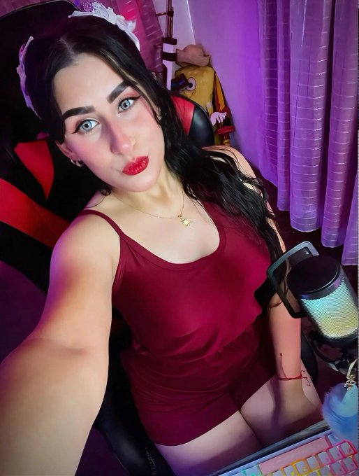 A woman in a red dress with a bow headband takes a selfie in her streaming setup, featuring a pink-lit gaming chair and a microphone. The image aligns with her post about starting the day with positive energy and streaming Axie Infinity on her Twitch and TikTok channels.