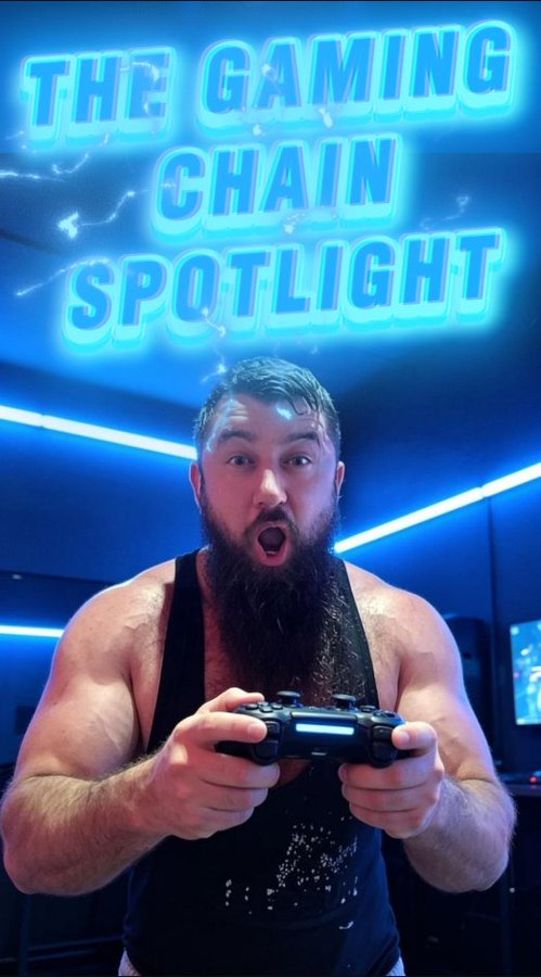 The image features a man with a beard holding a video game controller, possibly a Wii remote or a gamepad, as he appears to be playing a game. The background of the image has a blue neon sign that reads "The Gaming Chain Spotlight". This suggests that the man is participating in an event related to gaming or promoting a gaming-related business.