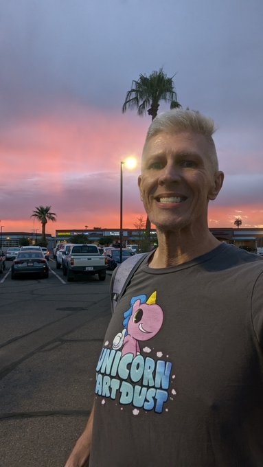 A smiling man with short blonde hair takes a selfie in a parking lot at sunset, wearing a gray t-shirt that reads "UNICORN FART DUST" with a pink unicorn graphic.