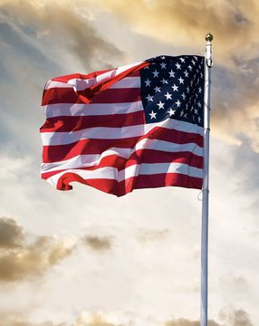 The image shows an American flag waving on a flagpole against a cloudy sky. The flag symbolizes the United States, directly relating to the announcement about Kraken expanding its onchain perpetual futures trading services to U.S. users.