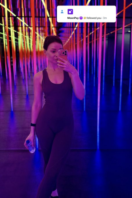 A mirror selfie of a woman in a black athletic outfit holding a phone and a water bottle in a room with colorful neon light installations, with a notification at the top showing 'MoonPay followed you'.