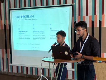 The image shows two men standing next to a laptop computer at an event where they are presenting their project. They appear to be using microphones for their presentation. The setting appears to be indoors, with a projector screen visible behind them. A whiteboard is also present in the scene.