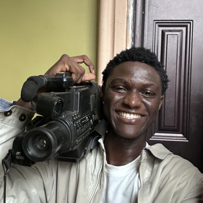 A smiling young man with short dark hair holds a professional Sony camcorder over his shoulder, looking directly at the camera.