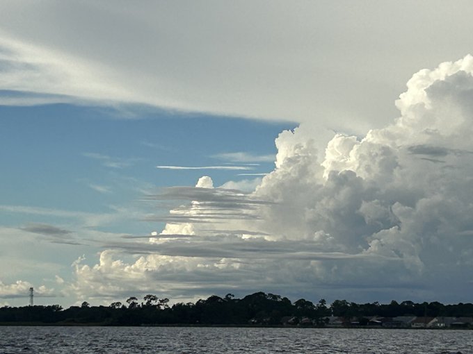 A scenic view of a dramatic sky with layered clouds over a dark treeline and water, captured near sunset.